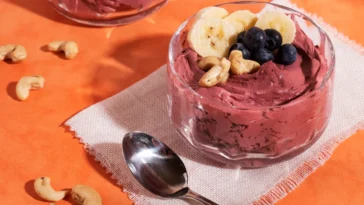 Tigela de sobremesa gelada com açaí cremoso, decorada com rodelas de banana, mirtilos frescos e castanhas-de-caju, servida sobre um guardanapo com uma colher ao lado.