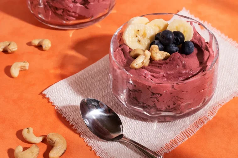 Tigela de sobremesa gelada com açaí cremoso, decorada com rodelas de banana, mirtilos frescos e castanhas-de-caju, servida sobre um guardanapo com uma colher ao lado.