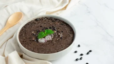 Tigela de sopa de feijão-preto decorada com folhas de hortelã e fatias de cebola roxa, acompanhada por colher de madeira e feijões espalhados em um fundo claro.