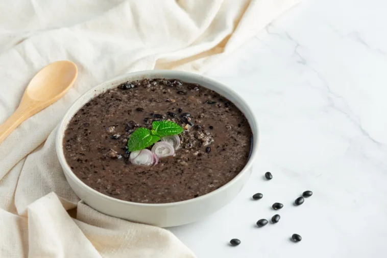 Tigela de sopa de feijão-preto decorada com folhas de hortelã e fatias de cebola roxa, acompanhada por colher de madeira e feijões espalhados em um fundo claro.