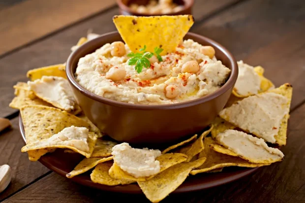 Bowl of hummus decorated with chickpeas, paprika and olive oil, surrounded by crunchy nachos arranged on a plate.