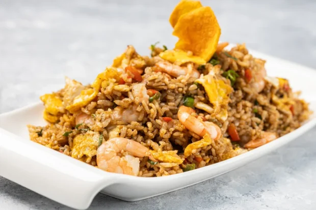 Prato de arroz frito com camarão, pedaços de omelete, vegetais e chips crocantes, servido em uma travessa branca sobre fundo cinza.