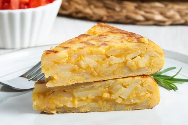 Duas fatias de tortilla espanhola com camadas de batata e ovos, dispostas em um prato branco com um ramo de alecrim e uma salada ao fundo.