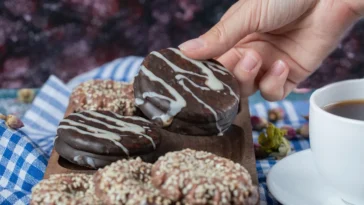 Mão segurando um pão de mel coberto com chocolate e decorado com fios de chocolate branco, acompanhado de outros biscoitos em uma tábua de madeira e uma xícara de café.