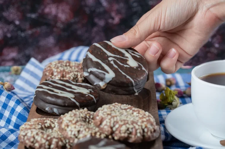 Mão segurando um pão de mel coberto com chocolate e decorado com fios de chocolate branco, acompanhado de outros biscoitos em uma tábua de madeira e uma xícara de café.