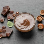 Tigela com creme de avelã cremoso, cercada por pedaços de chocolate com avelãs e avelãs inteiras sobre uma superfície escura.