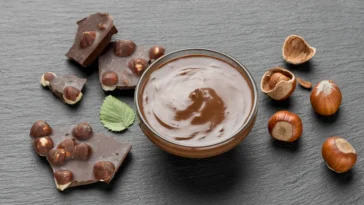 Tigela com creme de avelã cremoso, cercada por pedaços de chocolate com avelãs e avelãs inteiras sobre uma superfície escura.