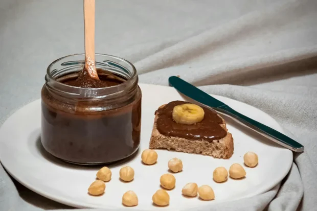 Pote de vidro com creme de avelã caseiro e colher mergulhada, acompanhado por uma fatia de pão integral com creme de avelã e banana, cercado por avelãs em um prato branco.