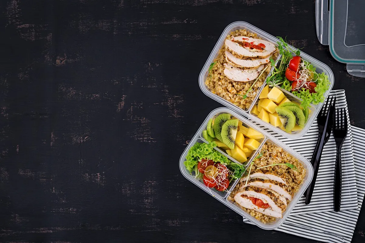Duas marmitas fitness organizadas com frango grelhado, arroz integral, salada e frutas como kiwi e manga, sobre uma superfície preta.