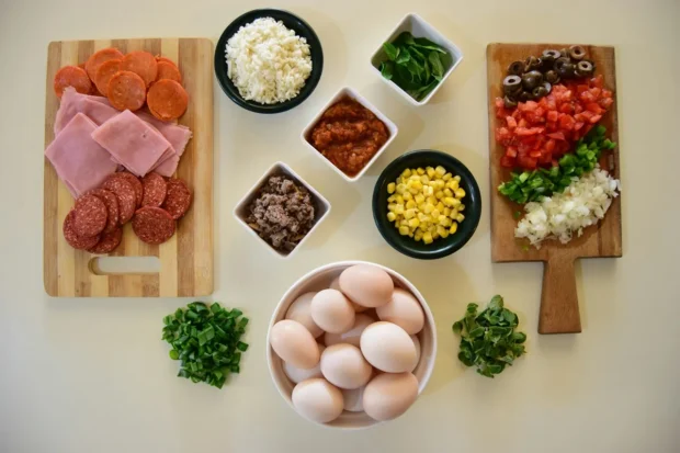 Mesa com ingredientes separados para preparo de receita, incluindo ovos, embutidos, milho, tomate, cebola, arroz, queijo, ervas e temperos.