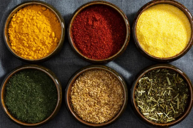 Seis tigelas de cerâmica contendo diferentes tipos de temperos e especiarias, como cúrcuma, páprica, fubá, ervas secas e sementes