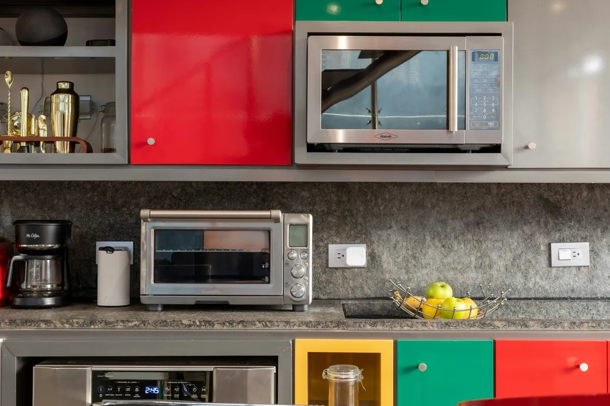 Cozinha moderna com armários coloridos, micro-ondas, forno elétrico, cafeteira e fruteira com maçãs sobre bancada de granito.